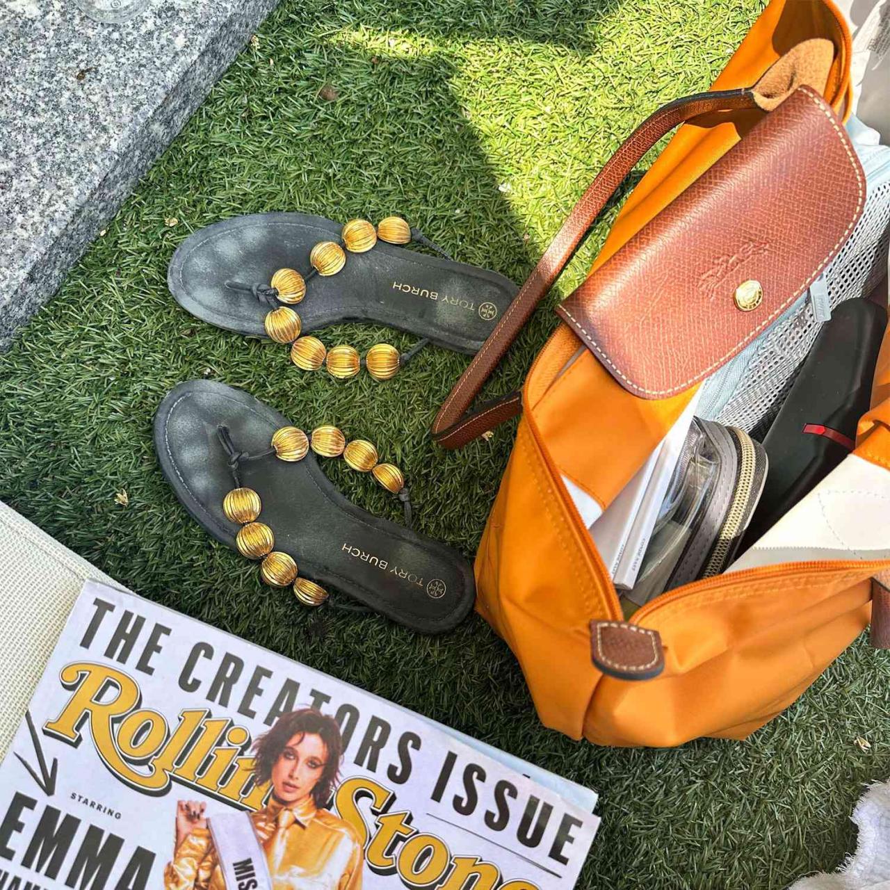 An orange Longchamp tote on the ground with Tory Burch sandals and Rolling Stone magazine.